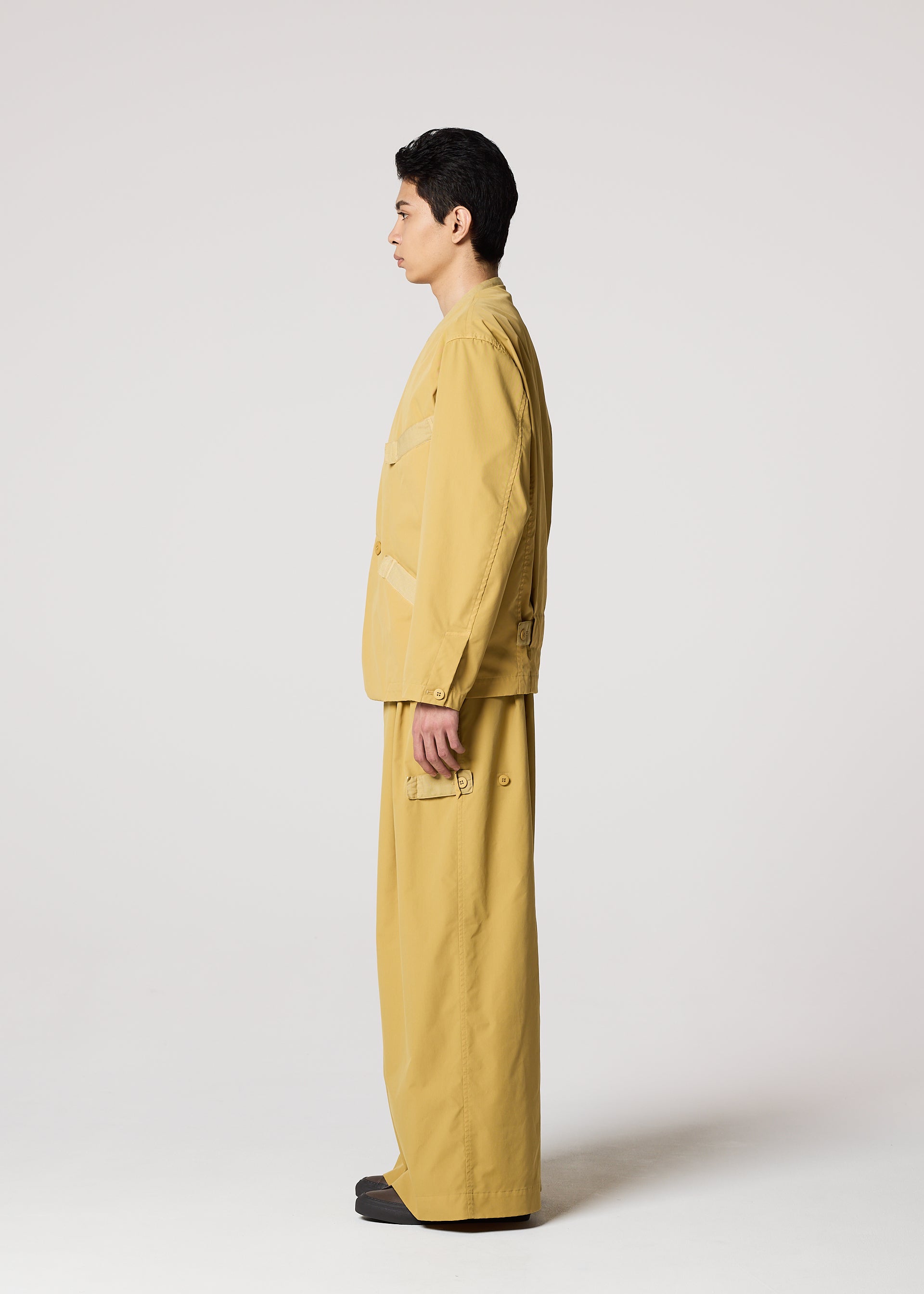 Zoomed view A person stands in profile facing left, wearing IM MEN's RESILIENCE set—a loose-fitting tan jacket and matching lightweight, wrinkle-resistant pants with pockets—perfect for everyday wear against a plain light background.