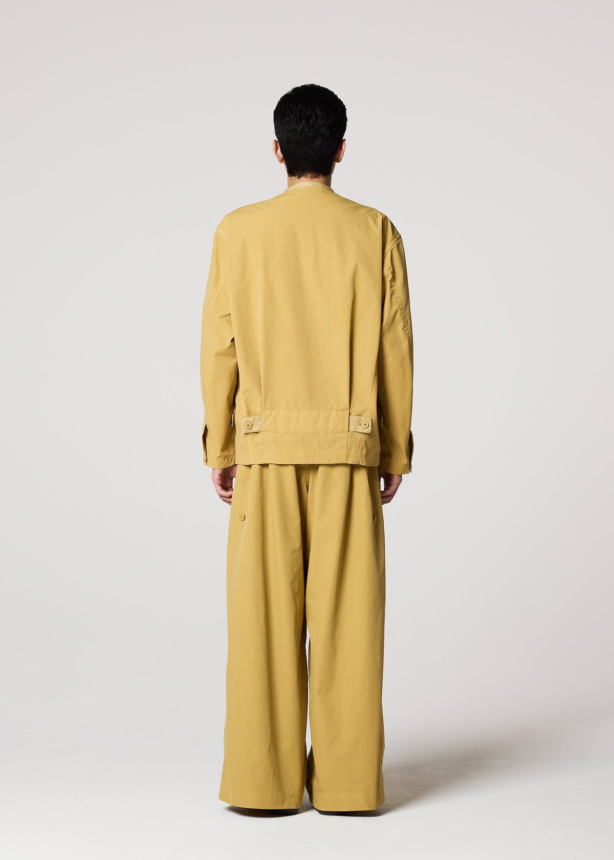 A person faces away from the camera, dressed in IM MEN's RESILIENCE: a tan, loose-fitting jacket with matching lightweight, wrinkle-resistant pants, set against a plain light background. view 8 of 9
