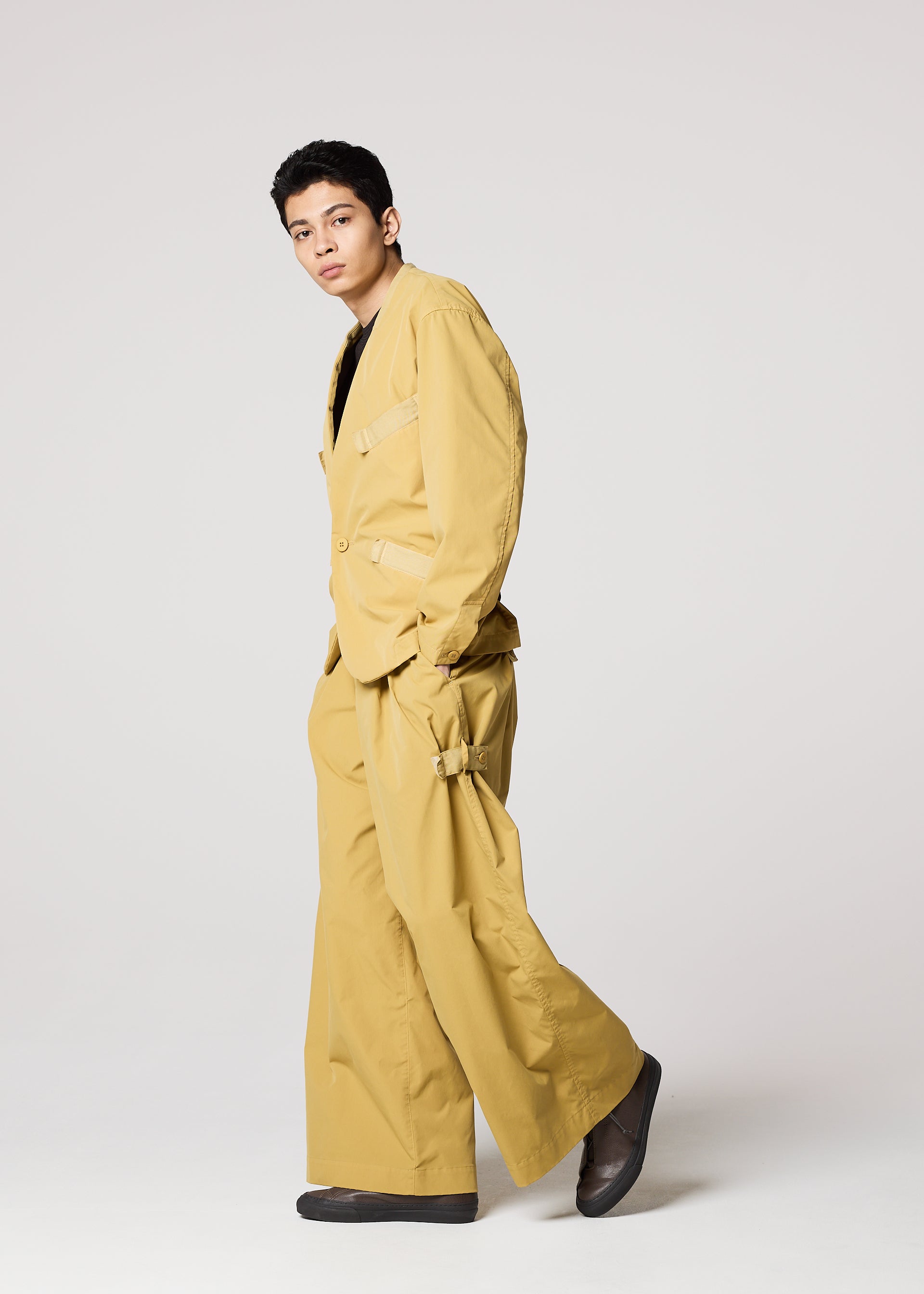 Zoomed view A person models the IM MEN RESILIENCE suit—a loose-fitting mustard yellow ensemble with wide-leg, lightweight, wrinkle-resistant pants and black shoes—posing confidently against a plain light background.