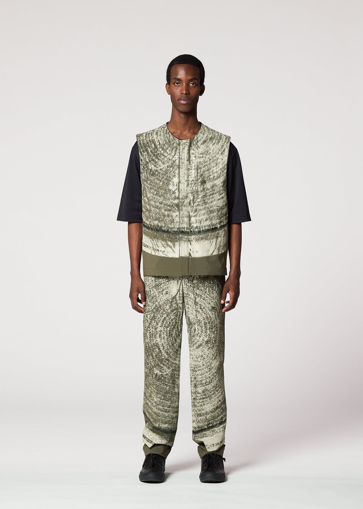 A man stands facing forward in the IM MEN KAIYU set—green and beige ceramic-inspired vest and ash glaze pants—over a black shirt with black shoes, against a plain white background. view 5 of 8