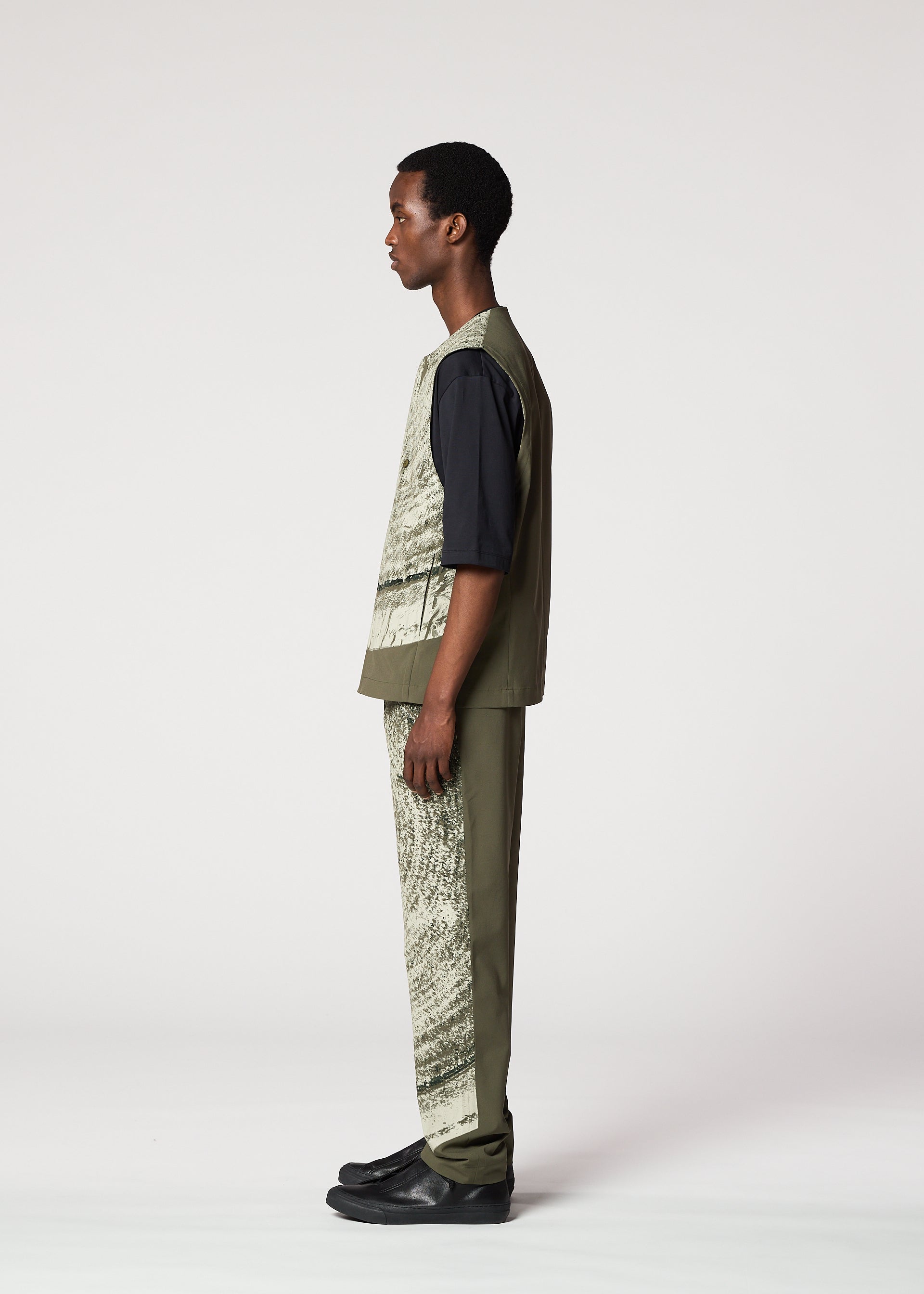 Zoomed view A person in profile poses against a plain background, wearing the IM MEN KAIYU set: a short-sleeved black shirt, ceramic-inspired green patterned vest and pigment print pants, paired with black shoes.