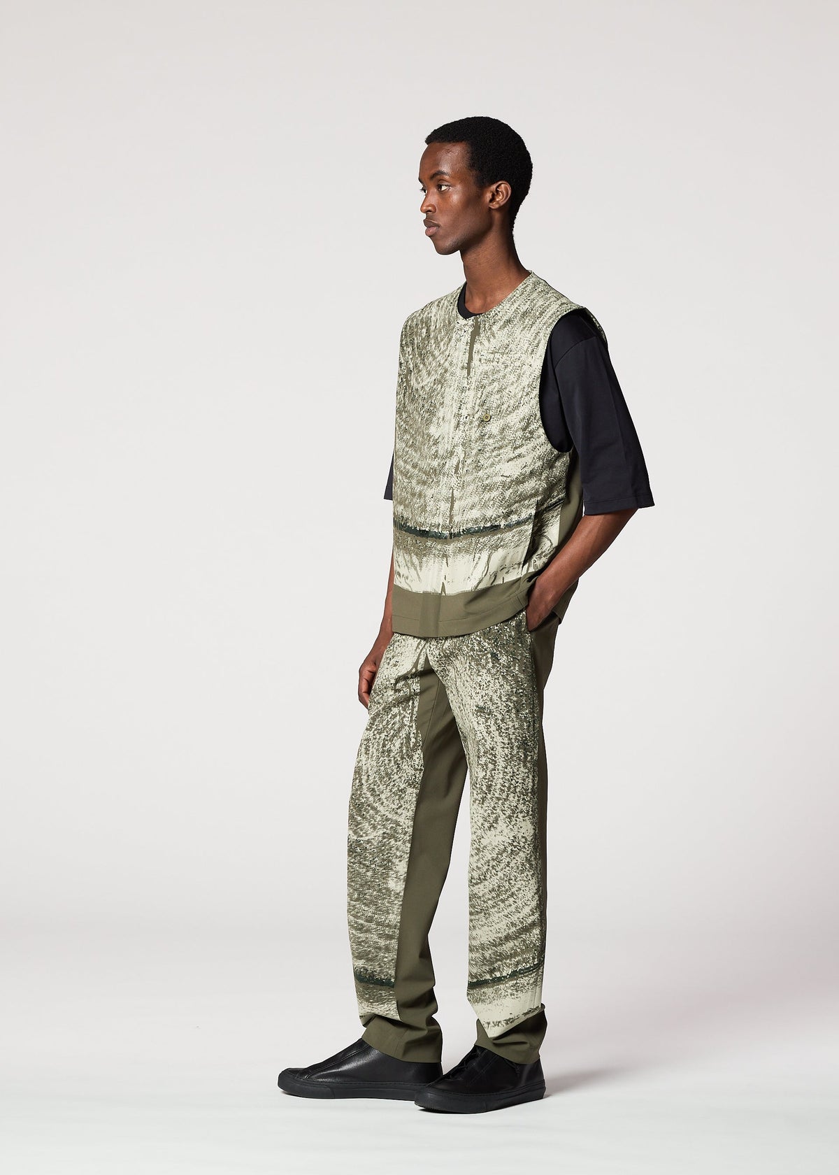 A man models the IM MEN KAIYU sleeveless patterned vest over a black shirt and ash glaze pants with green panels, paired with black shoes. He stands in a studio, facing slightly right against a plain background. view 8 of 8