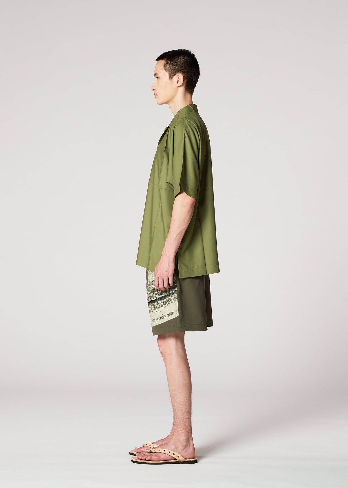 A man stands in profile against a plain light background, wearing the IM MEN KAIYU olive green short-sleeve shirt, matching pigment-printed shorts, and beige flip-flops. view 6 of 8