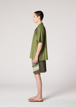 A man stands in profile against a plain light background, wearing the IM MEN KAIYU olive green short-sleeve shirt, matching pigment-printed shorts, and beige flip-flops.