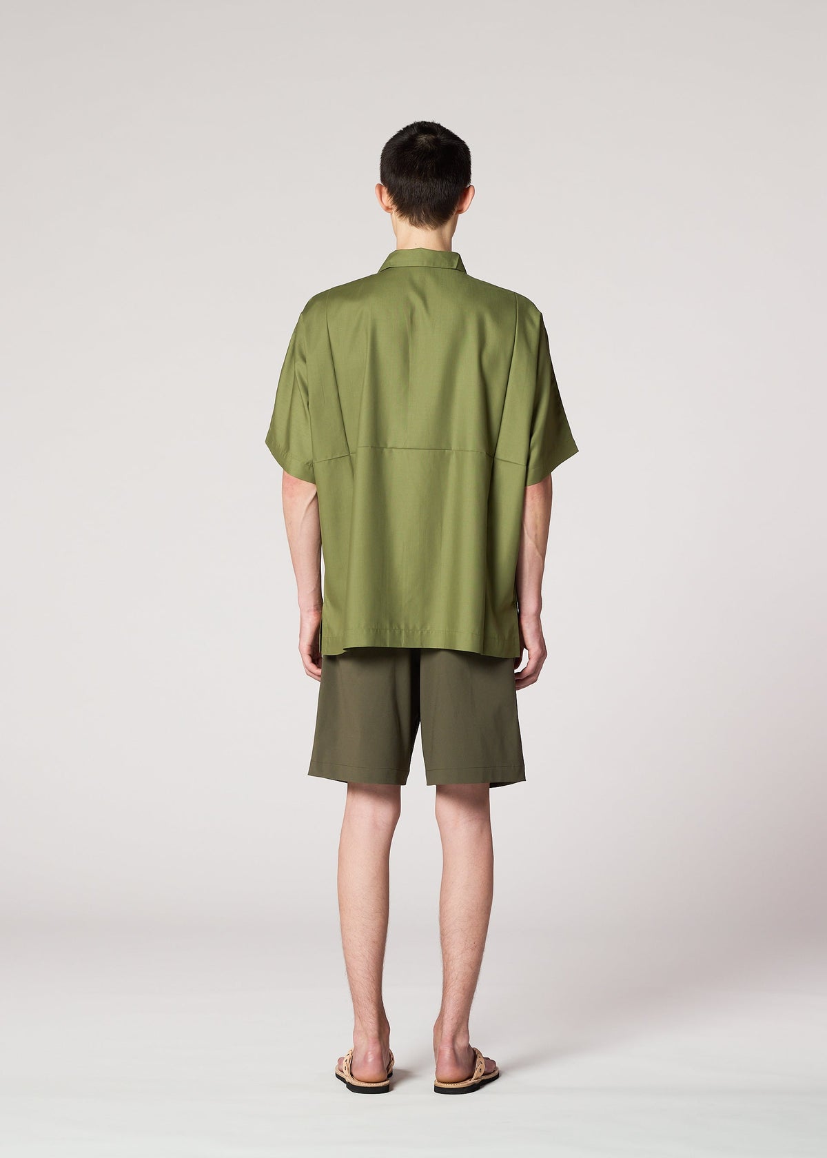 Someone stands with their back to the camera, wearing IM MEN's KAIYU olive green short-sleeve shirt and celadon shorts, paired with sandals, against a plain light background. view 7 of 8