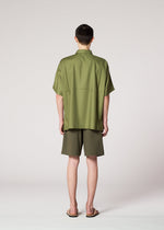 Someone stands with their back to the camera, wearing IM MEN's KAIYU olive green short-sleeve shirt and celadon shorts, paired with sandals, against a plain light background.