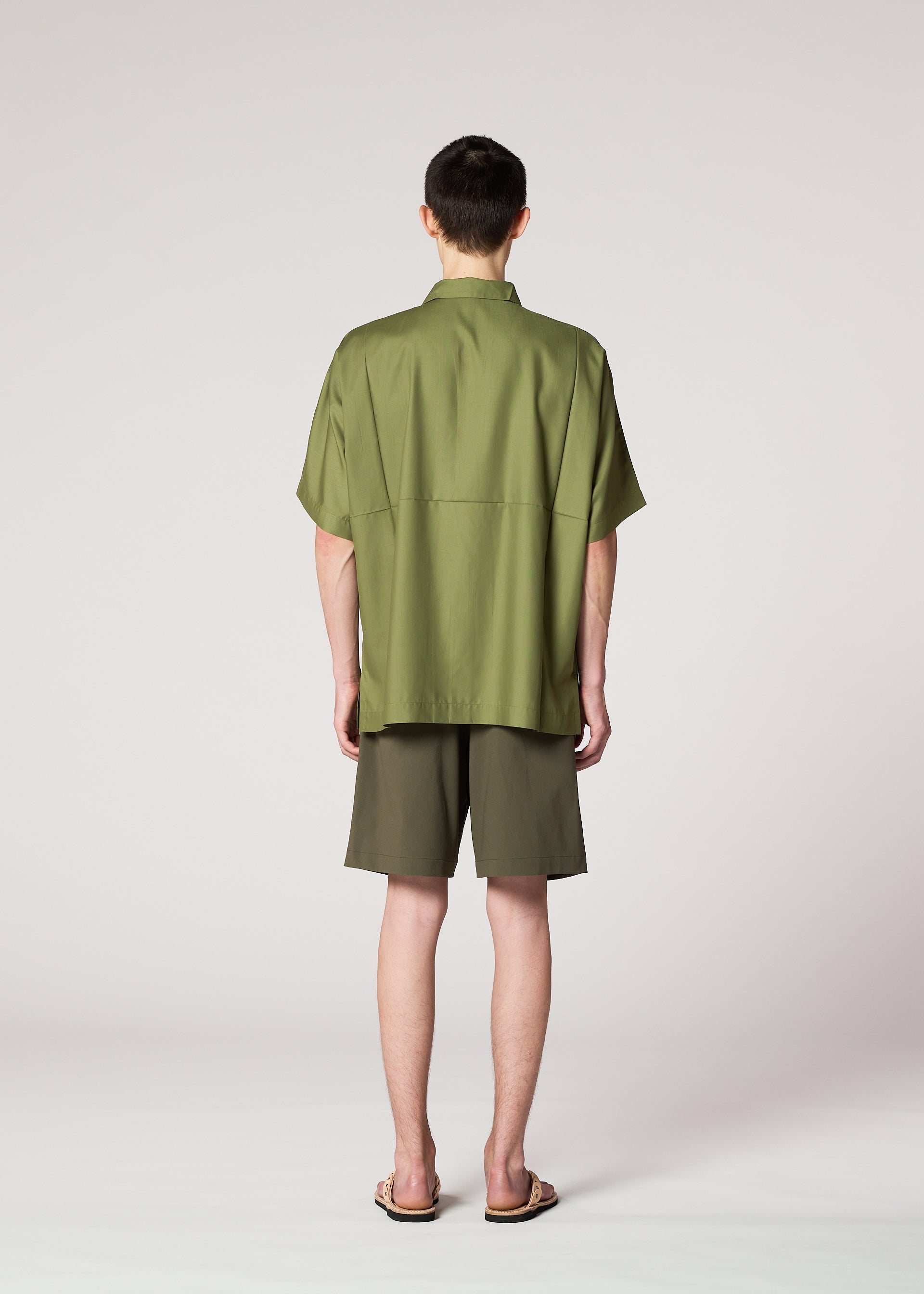 Zoomed view Someone stands with their back to the camera, wearing IM MEN's KAIYU olive green short-sleeve shirt and celadon shorts, paired with sandals, against a plain light background.