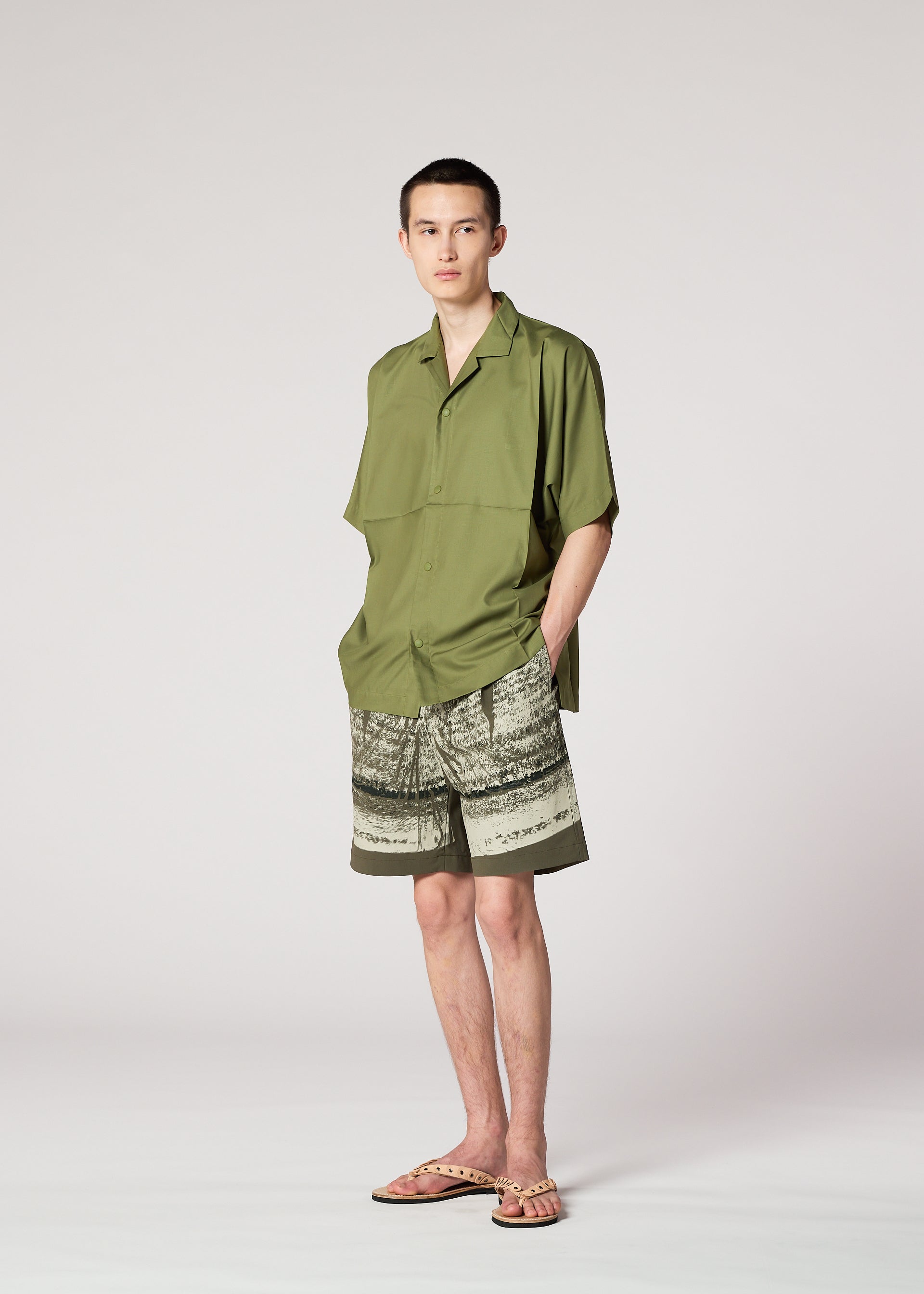 Zoomed view A person with a neutral expression stands against a plain background wearing the IM MEN KAIYU olive green short-sleeve shirt, ceramic-inspired pigment-printed green and beige shorts, and beige sandals, with hands in pockets.