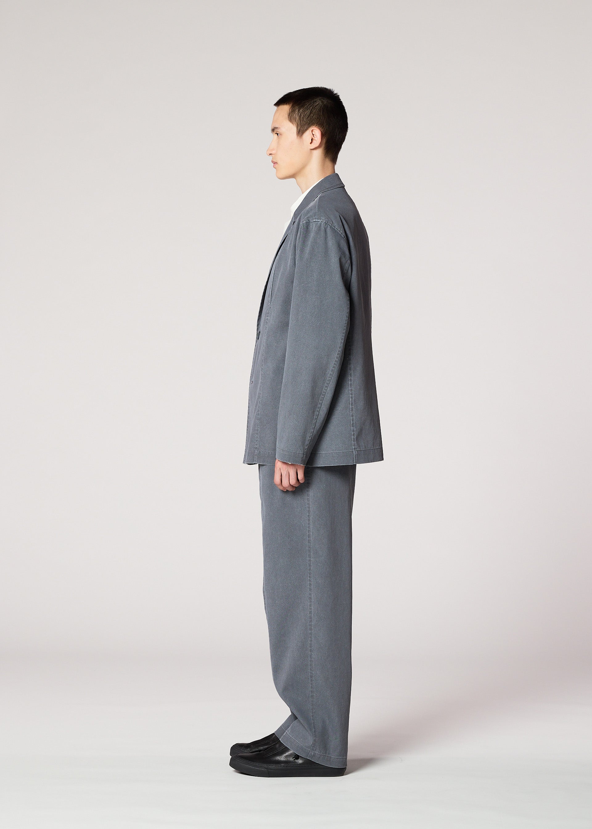 Zoomed view A man stands in profile against a plain background, wearing the IM MEN AIR WASH loose-fitting gray suit with wide-legged, lightweight pants made from plant-derived polyester, paired with black shoes.