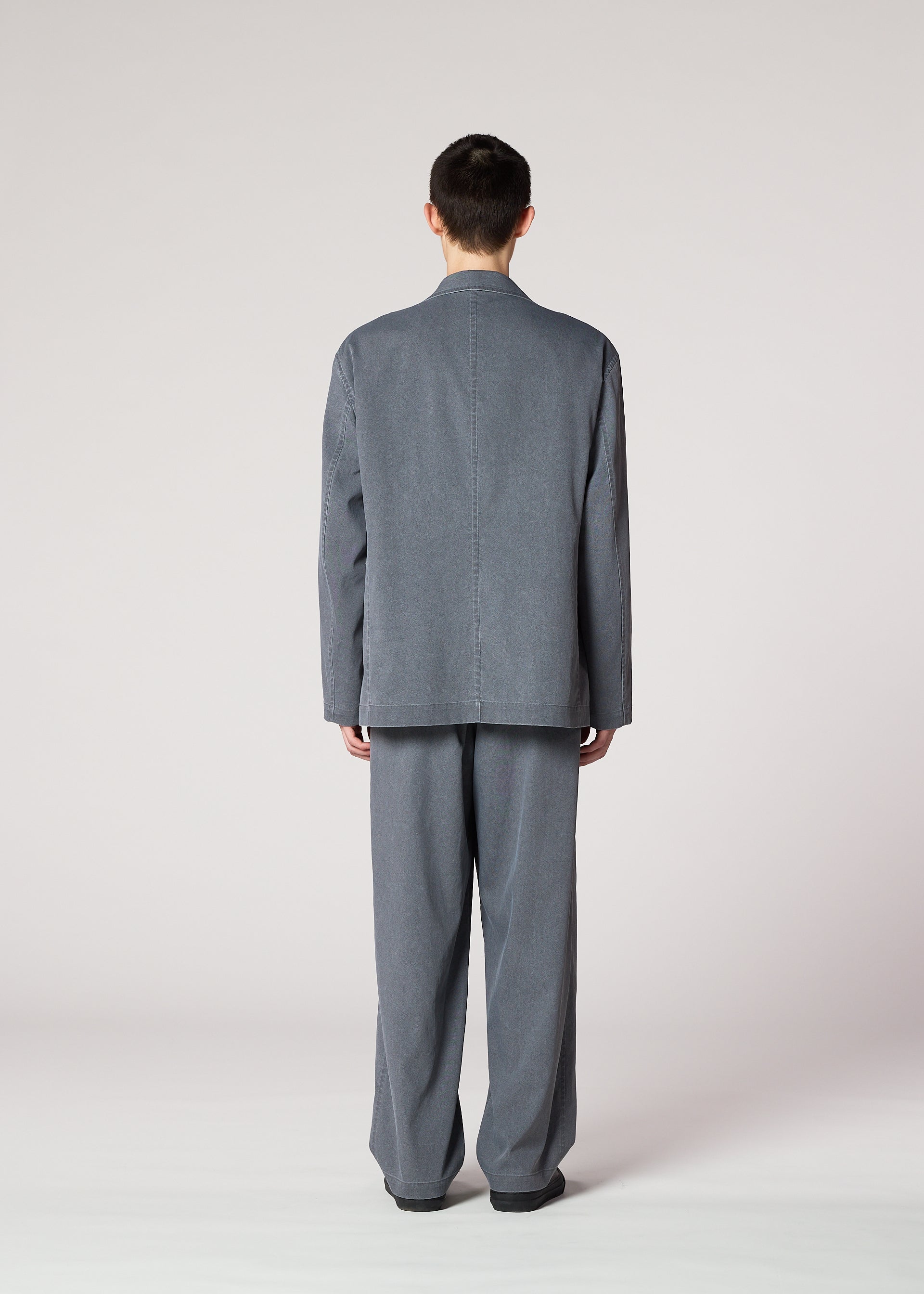 Zoomed view A person faces away from the camera, wearing IM MEN's AIR WASH: a gray, loose-fitting suit with long sleeves and lightweight pants made of plant-derived polyester, shown against a plain, light background.