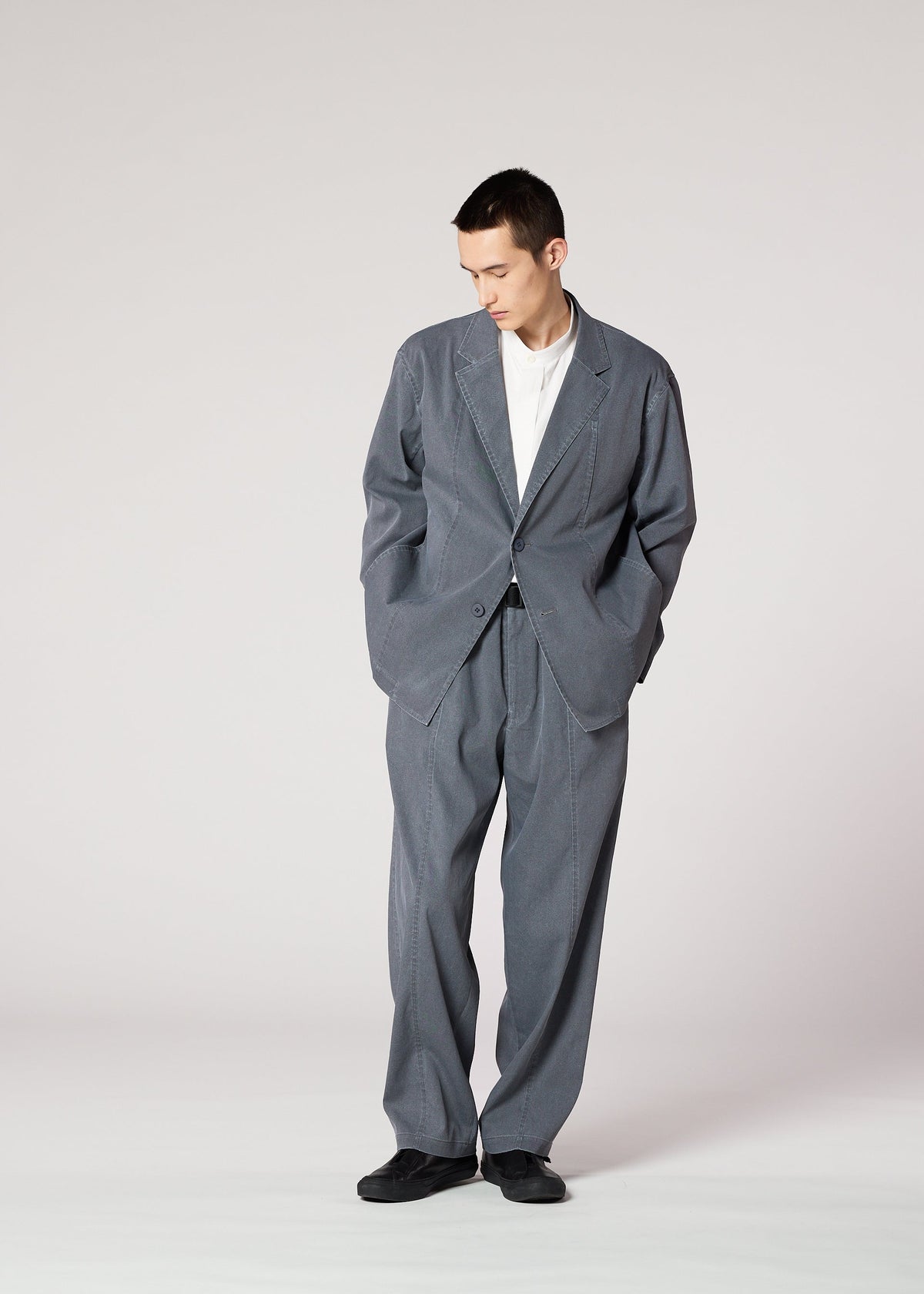 A person stands against a plain background, wearing IM MEN's AIR WASH lightweight, loose-fitting gray pants made from plant-derived polyester, paired with a white shirt and black shoes, looking down with hands in pockets. view 8 of 8