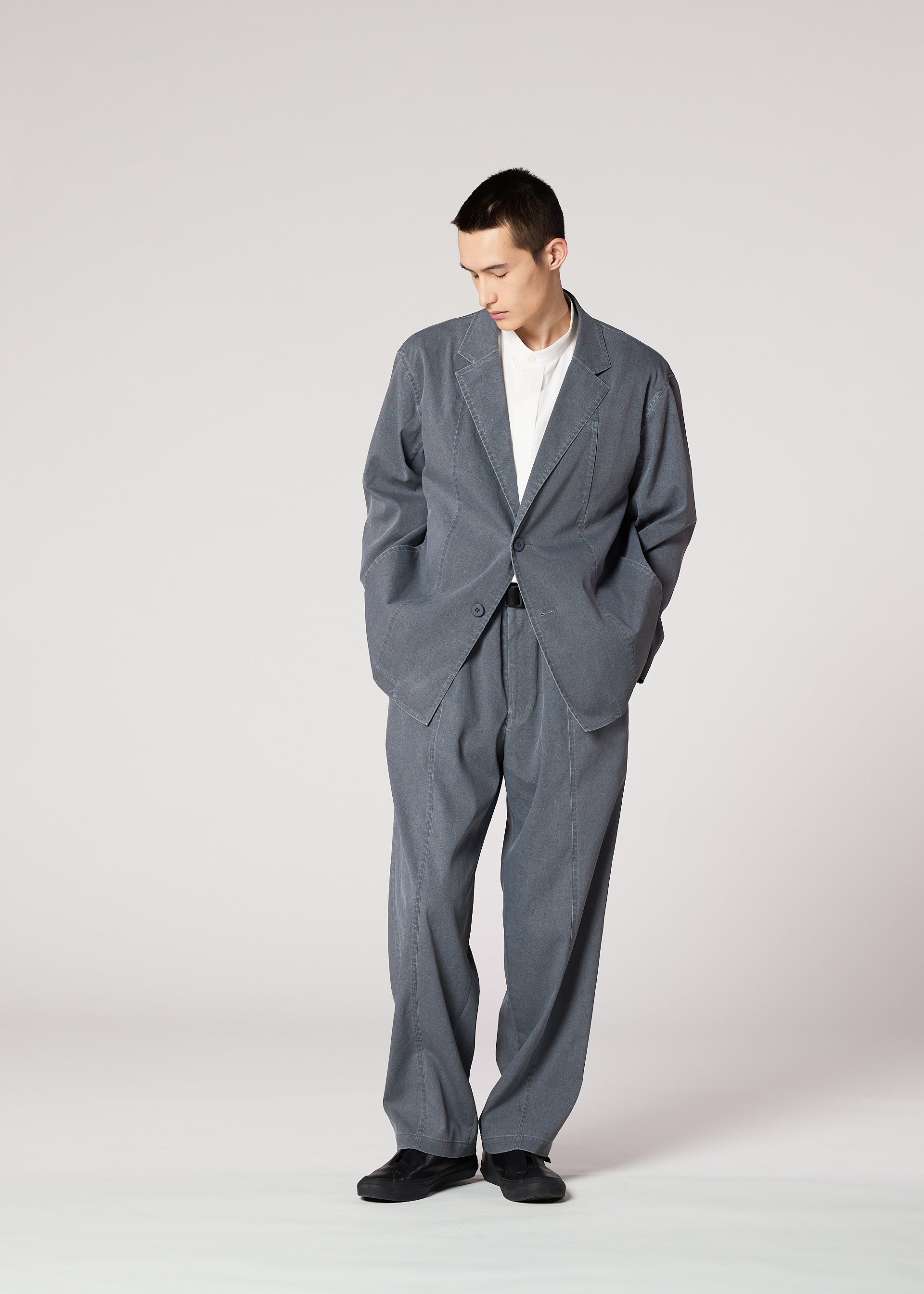 Zoomed view A person stands against a plain background, wearing IM MEN's AIR WASH lightweight, loose-fitting gray pants made from plant-derived polyester, paired with a white shirt and black shoes, looking down with hands in pockets.