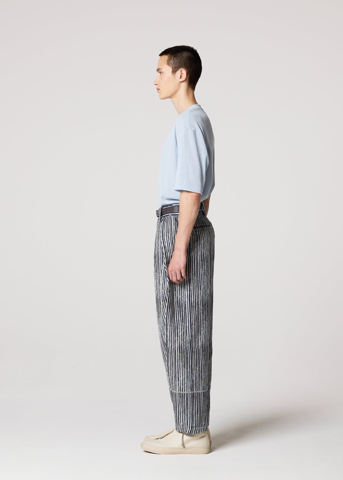 A person stands in profile against a plain light background, wearing the IM MEN CURVINESS light blue t-shirt and black and white striped cotton linen textured pants, paired with beige shoes. view 6 of 8