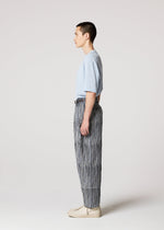 A person stands in profile against a plain light background, wearing the IM MEN CURVINESS light blue t-shirt and black and white striped cotton linen textured pants, paired with beige shoes.