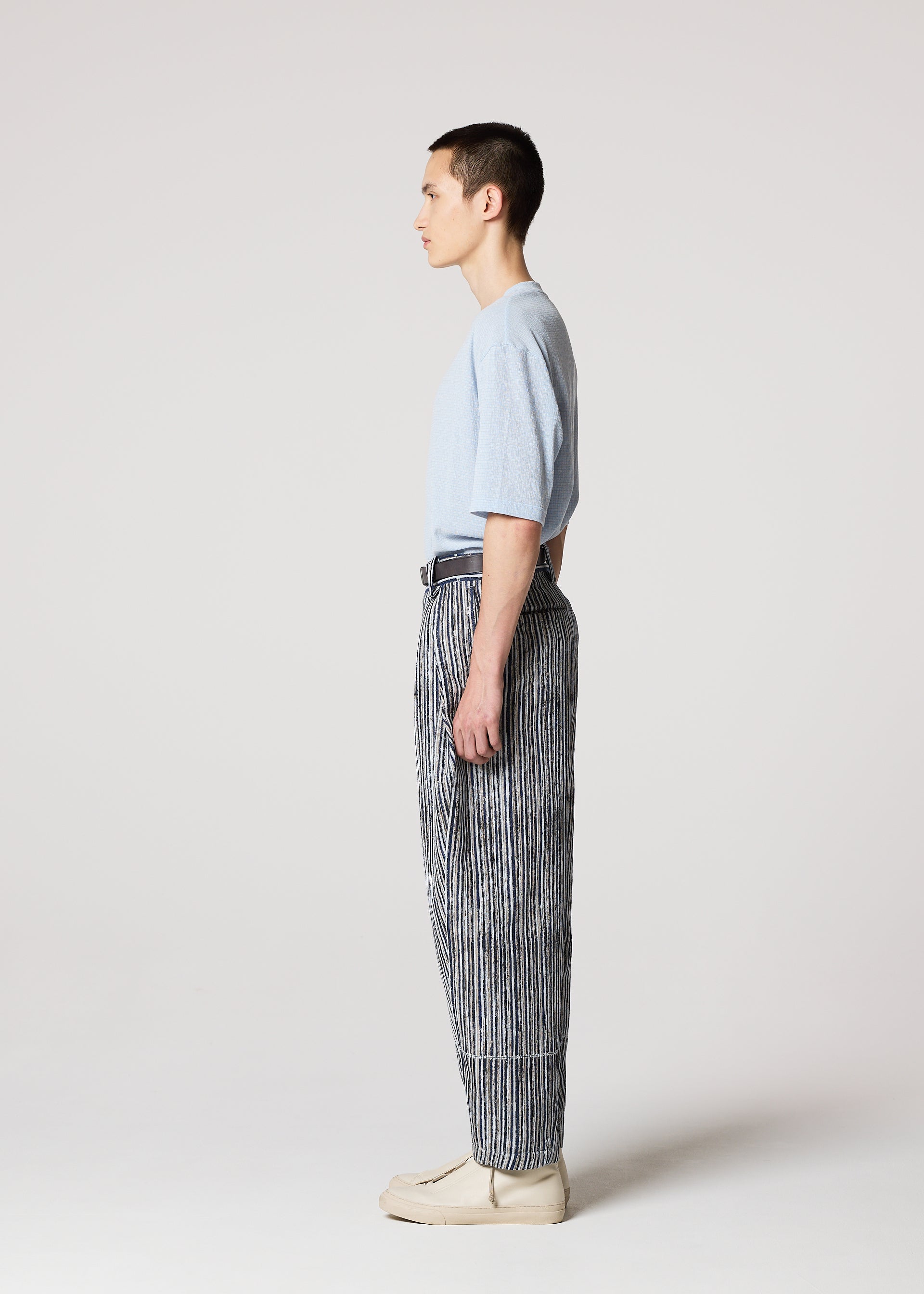 Zoomed view A person stands in profile against a plain light background, wearing the IM MEN CURVINESS light blue t-shirt and black and white striped cotton linen textured pants, paired with beige shoes.
