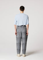 A person faces away from the camera, wearing the IM MEN CURVINESS light blue short-sleeve top and high-waisted ceramic artist-inspired black and white striped pants, paired with beige shoes against a plain light background.