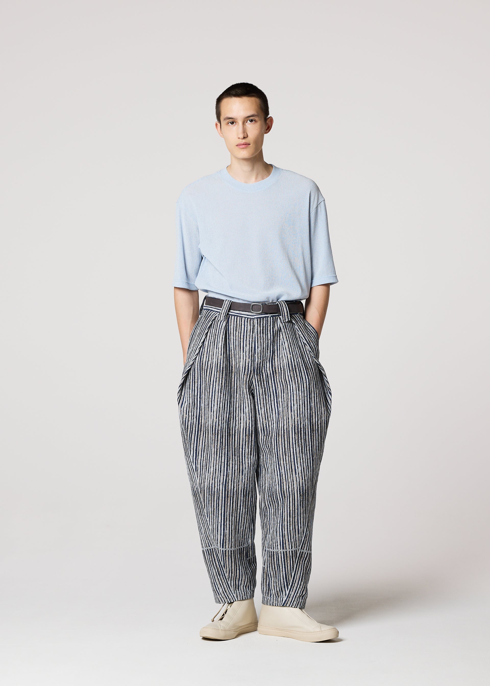 Zoomed view A person stands facing forward, wearing IM MEN's CURVINESS light blue t-shirt tucked into high-waisted, loose cotton-linen textured pants with a belt and light shoes against a plain, light background.