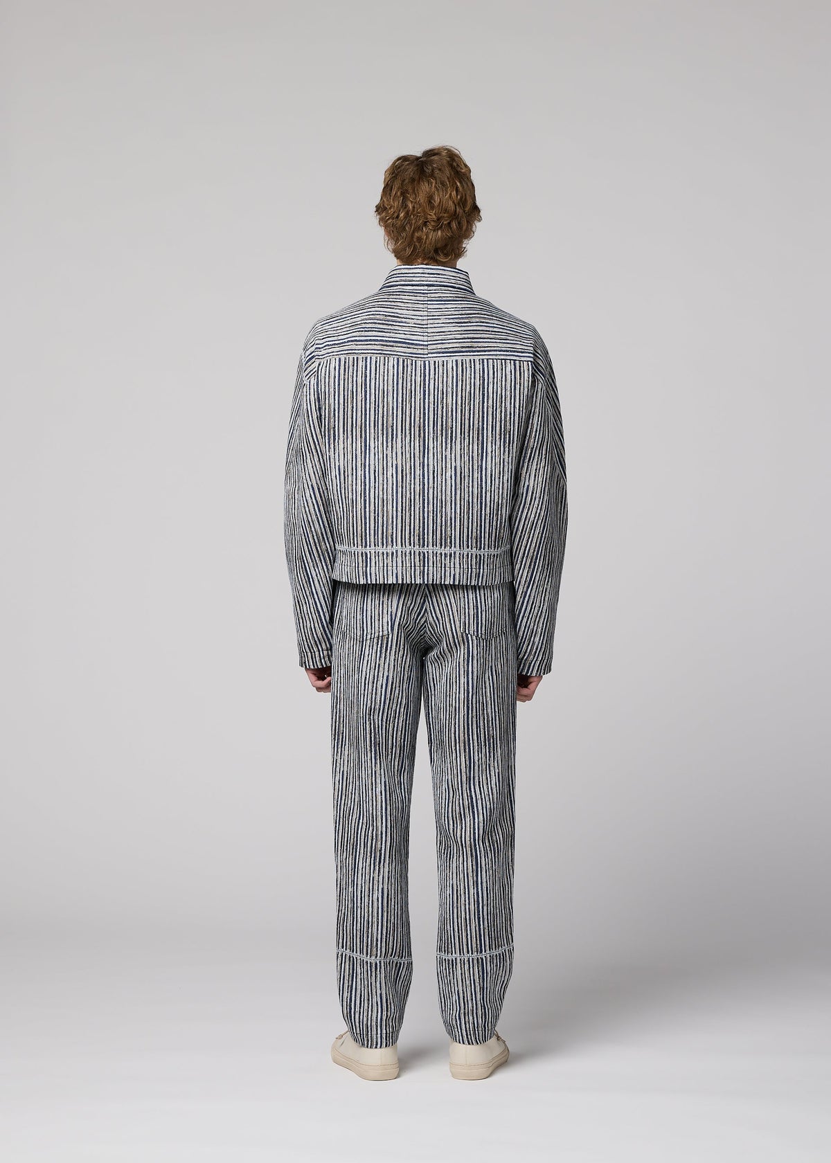 A person faces away wearing the CURVINESS blue and white jacquard loom pants and striped jacket by IM MEN with white shoes, set against a plain light gray background. view 6 of 7