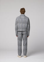 A person faces away wearing the CURVINESS blue and white jacquard loom pants and striped jacket by IM MEN with white shoes, set against a plain light gray background.
