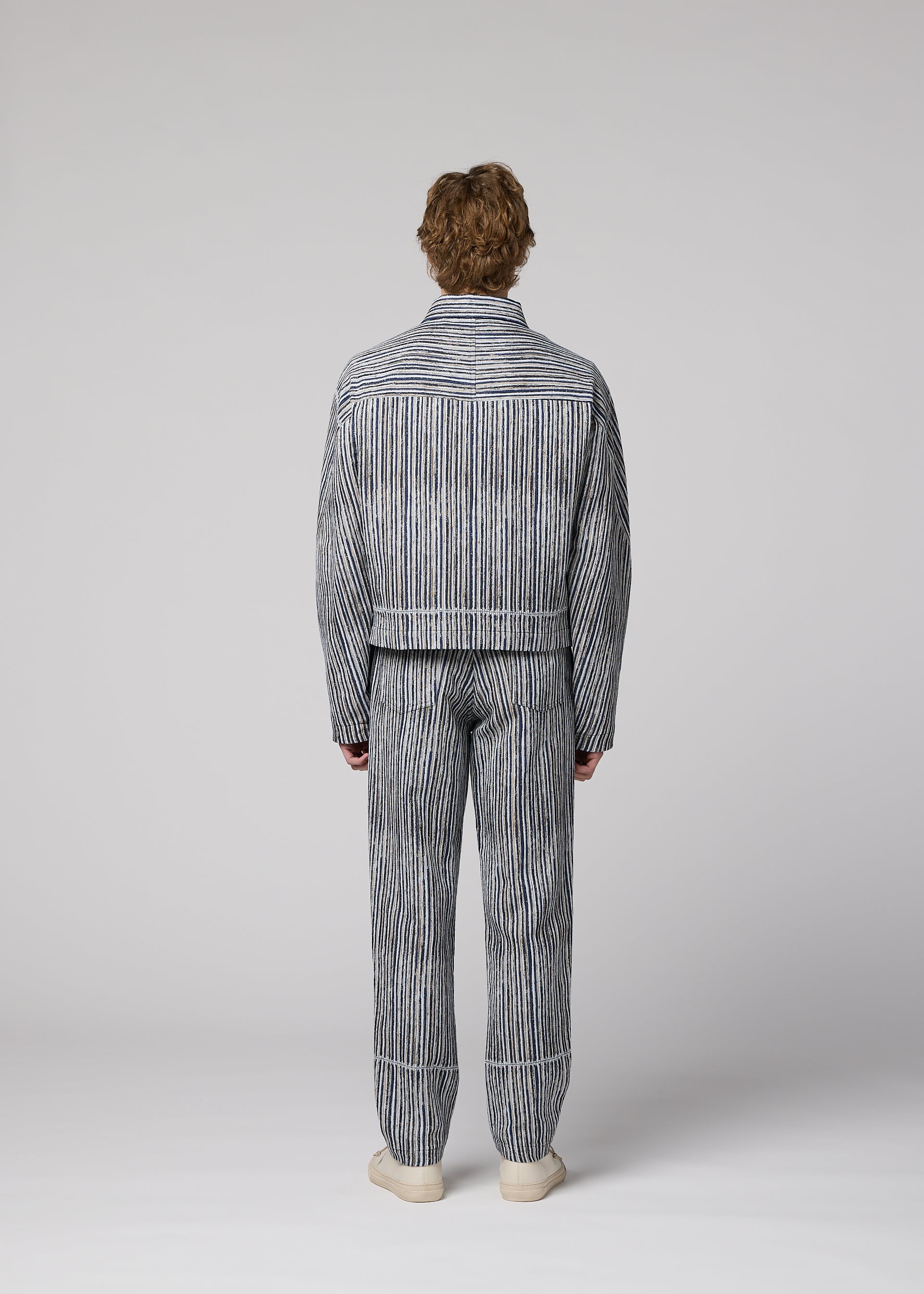 Zoomed view A person faces away wearing the CURVINESS blue and white jacquard loom pants and striped jacket by IM MEN with white shoes, set against a plain light gray background.