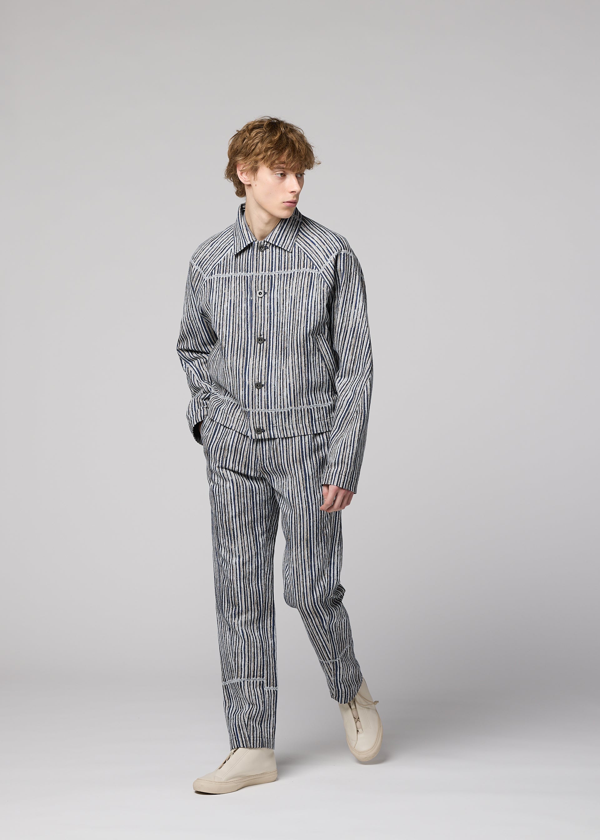 Zoomed view A person with wavy hair stands against a light gray background, wearing IM MEN's CURVINESS ceramic-inspired pants and a striped jacket, paired with white shoes, gazing slightly to the side.