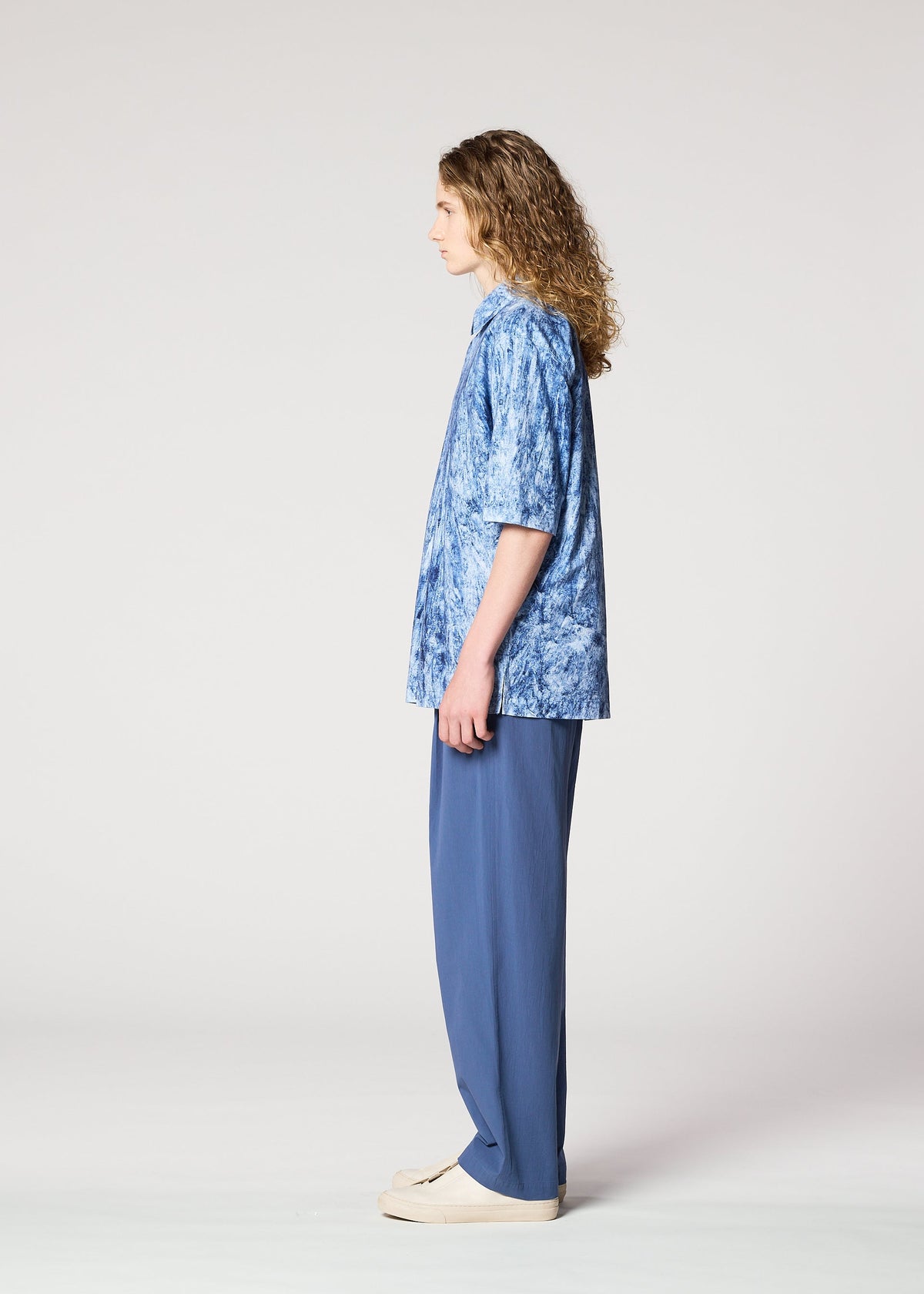A person with long curly hair stands in profile against a plain background, wearing a blue tie-dye short-sleeve shirt, IM MEN NAVY BOTTOMS, and light-colored shoes. view 6 of 8