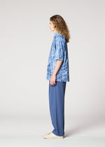 A person with long curly hair stands in profile against a plain background, wearing a blue tie-dye short-sleeve shirt, IM MEN NAVY BOTTOMS, and light-colored shoes.