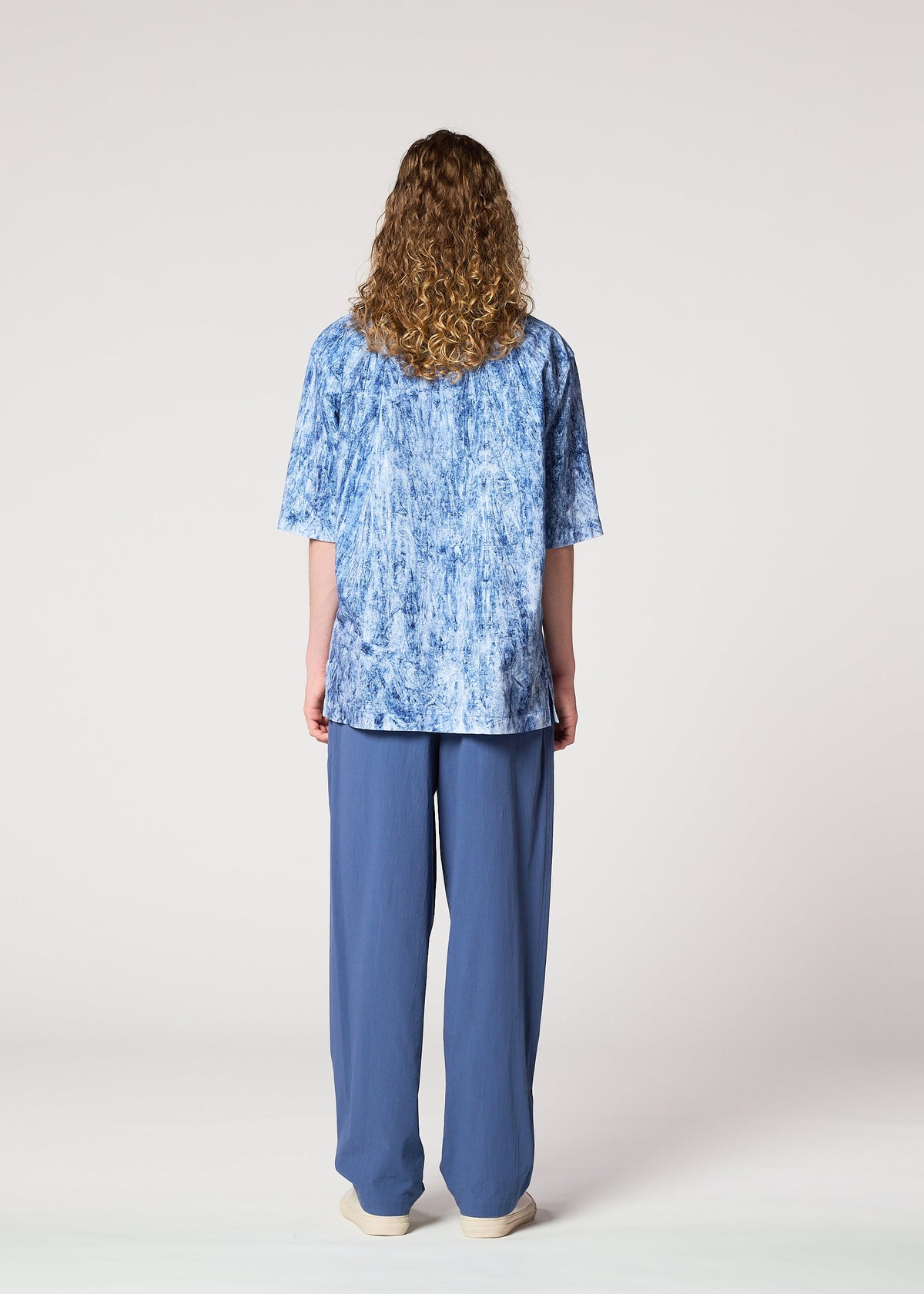 A person with long curly hair stands facing away, wearing a loose blue patterned shirt, IM MEN’s NAVY BOTTOMS, and beige shoes against a plain light background. view 7 of 8