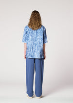 A person with long curly hair stands facing away, wearing a loose blue patterned shirt, IM MEN’s NAVY BOTTOMS, and beige shoes against a plain light background.