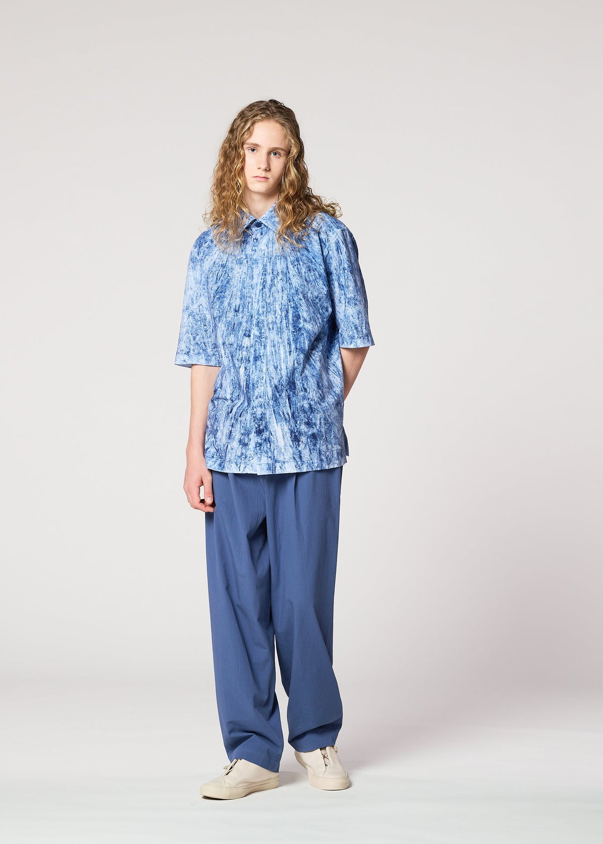 A person with long curly hair stands against a plain background, wearing a loose-fitting blue patterned short-sleeve shirt, IM MEN NAVY BOTTOMS, and white shoes. view 8 of 8