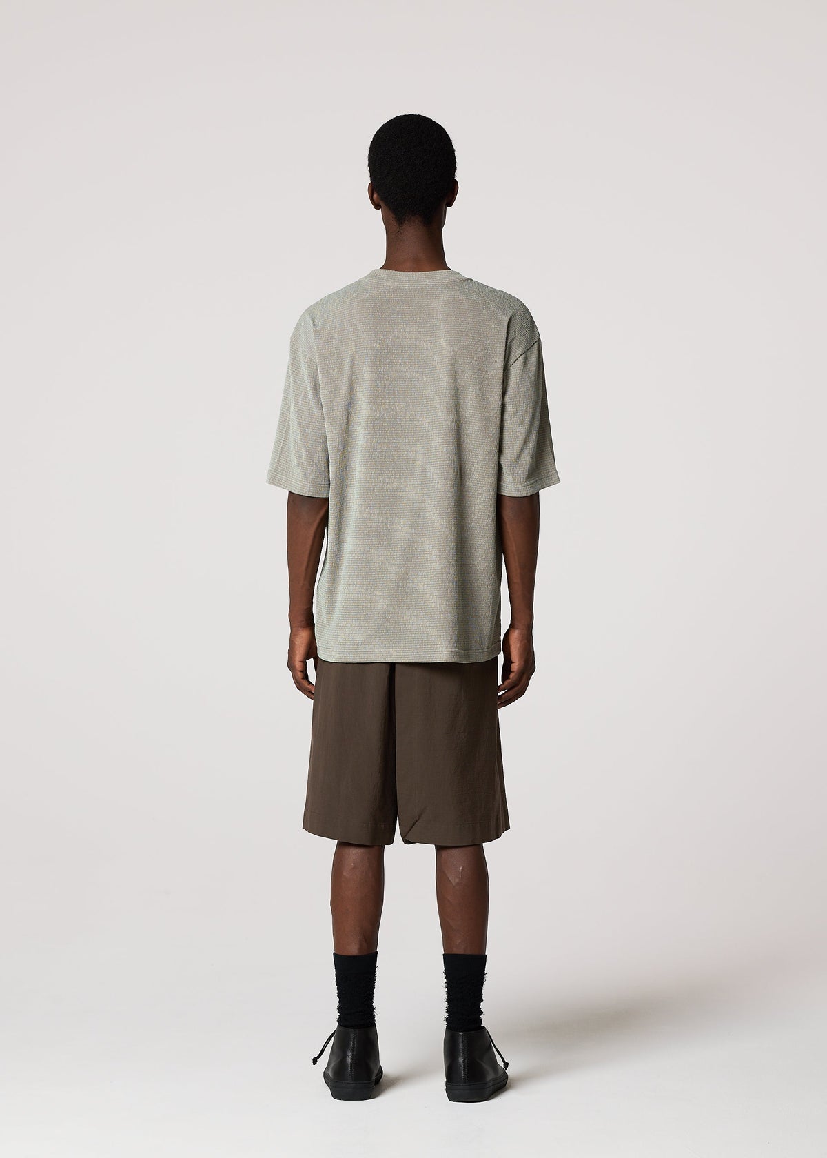 A person faces away from the camera, wearing a loose gray t-shirt, NAVY BOTTOMS by IM MEN, black socks, and black shoes against a plain white background. view 8 of 9