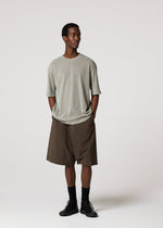 A man stands against a plain light background, wearing loose-fitting gray T-shirt and IM MEN’s NAVY BOTTOMS that fall to the knee. He pairs the look with black socks and shoes, hands in his pockets as he gazes off to the side.