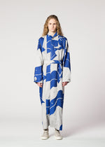 A person with long, wavy hair wears the INLAY dress by IM MEN—a long, belted piece made from recycled polyester in a bold blue and gray abstract pattern—standing against a plain white background with a neutral expression and white sneakers.