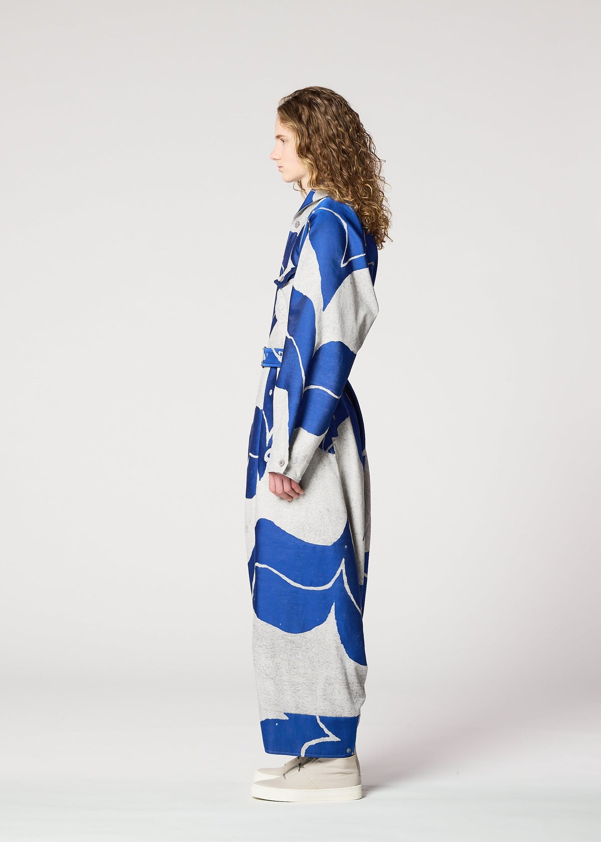 A person with curly hair stands in profile wearing IM MEN's INLAY long, belted coat made from recycled polyester in a bold blue and gray abstract print, paired with white platform shoes, against a plain light background. view 4 of 6