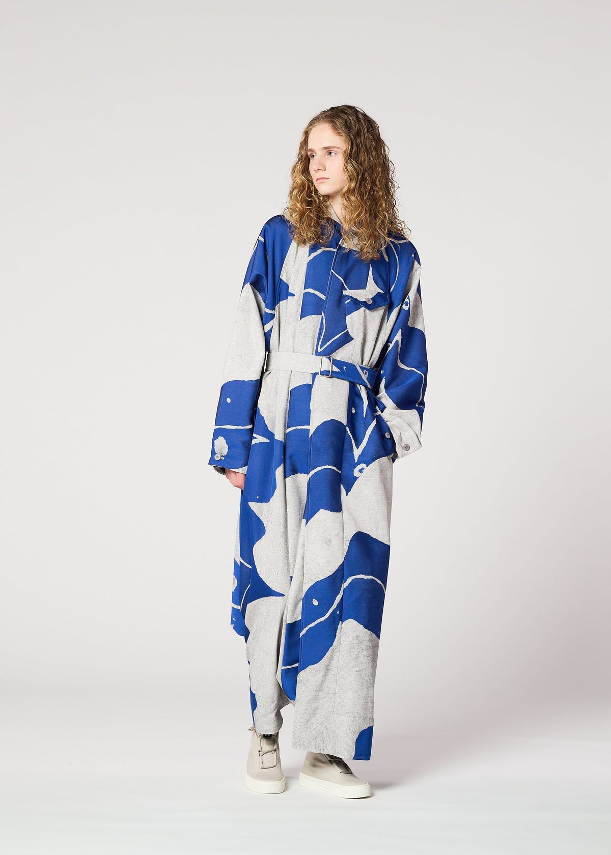 A person with long curly hair models the IM MEN INLAY blue and gray jacquard weave, loose-fitting dress with a belt, standing against a plain light background, gazing to the side with one hand in a pocket. view 6 of 6