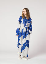 A person with long curly hair models the IM MEN INLAY blue and gray jacquard weave, loose-fitting dress with a belt, standing against a plain light background, gazing to the side with one hand in a pocket.