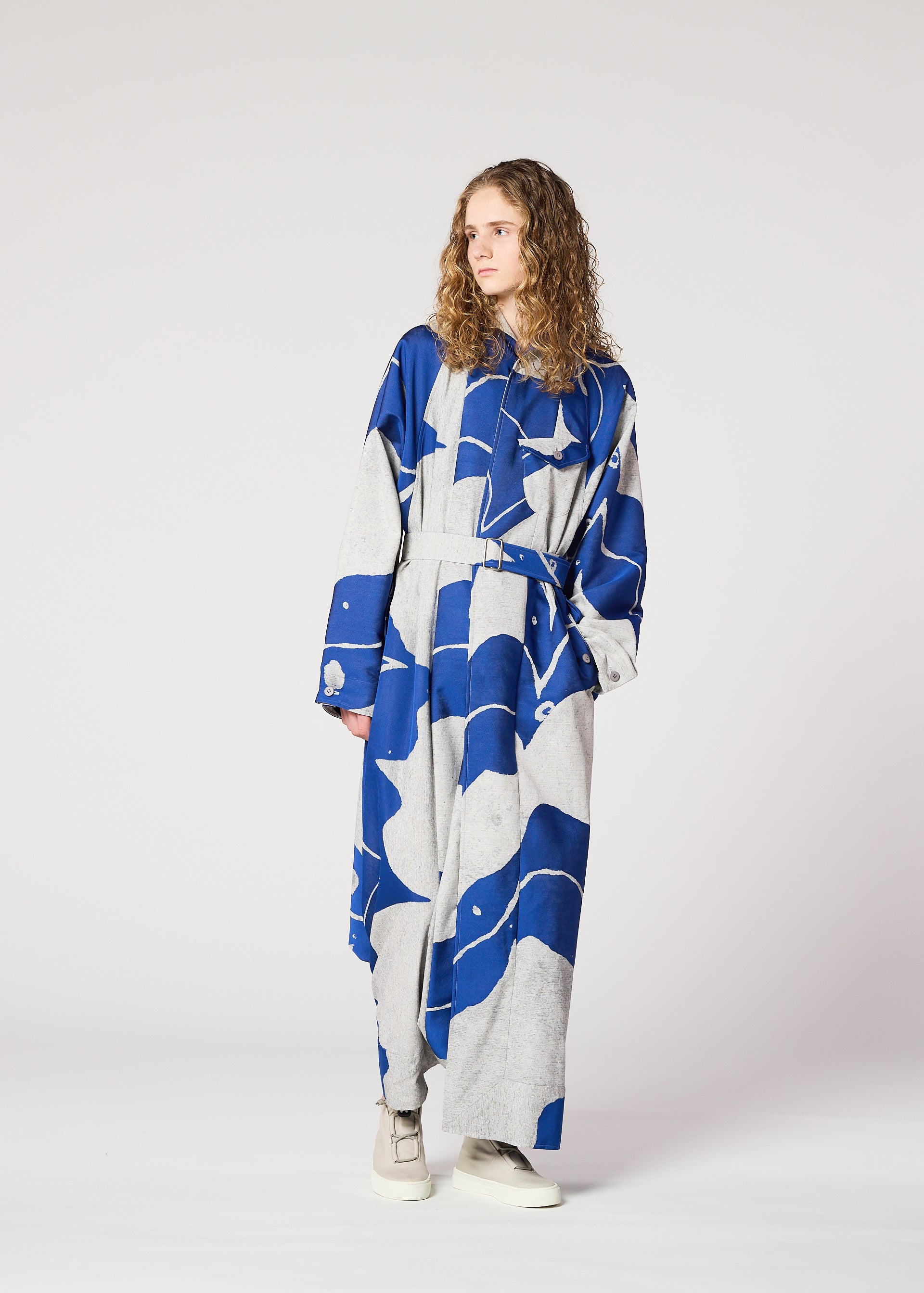 Zoomed view A person with long curly hair models the IM MEN INLAY blue and gray jacquard weave, loose-fitting dress with a belt, standing against a plain light background, gazing to the side with one hand in a pocket.