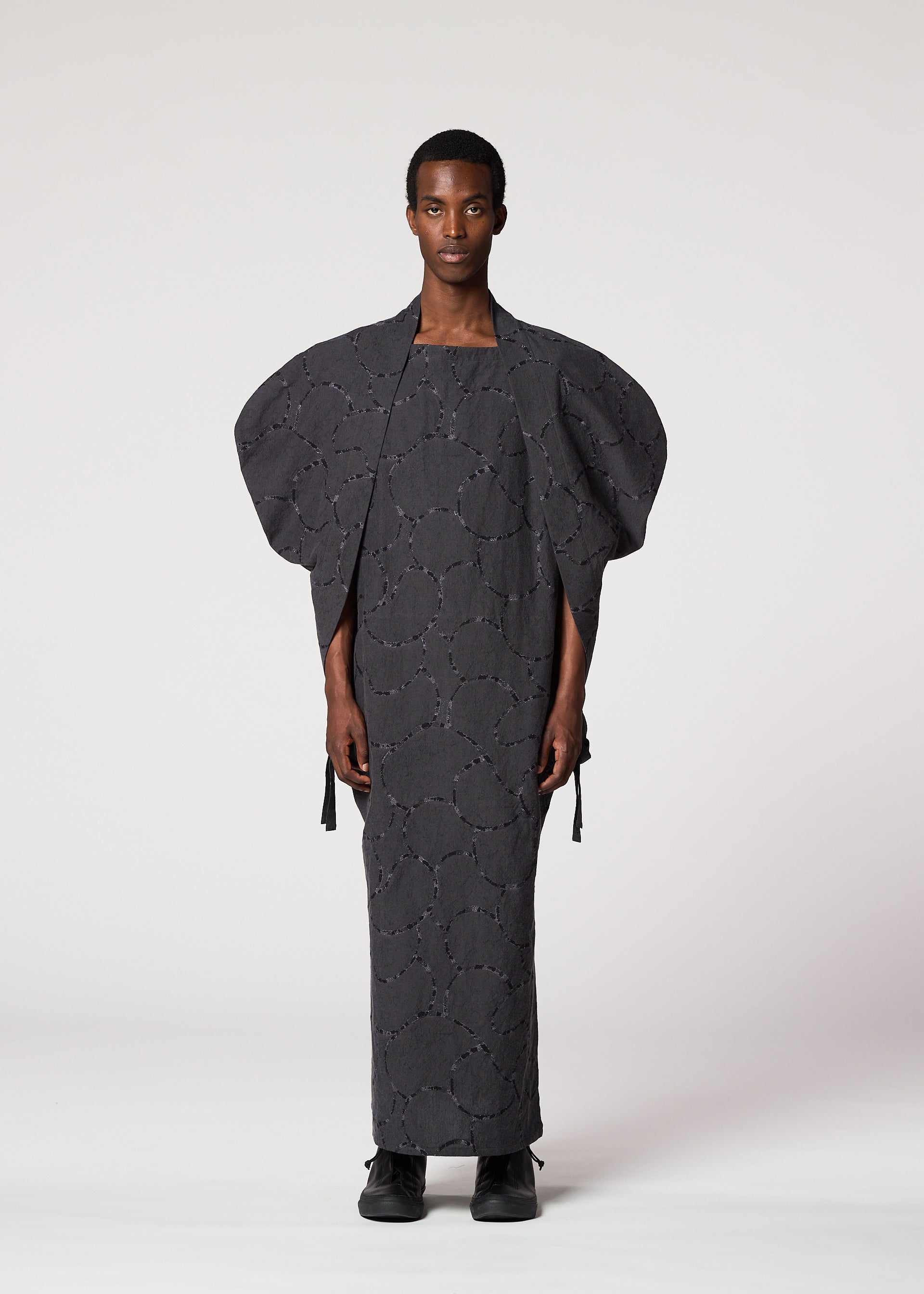 Zoomed view A person stands against a plain background, wearing the IM MEN VASE JACQUARD SHIRT with wide, structured sleeves and circular patterns. They have a calm expression and are wearing black shoes.