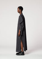 A person stands in profile wearing the IM MEN VASE JACQUARD SHIRT, a long, dark, textured piece with ruffled details and side slits, paired with black shoes against a plain white background.