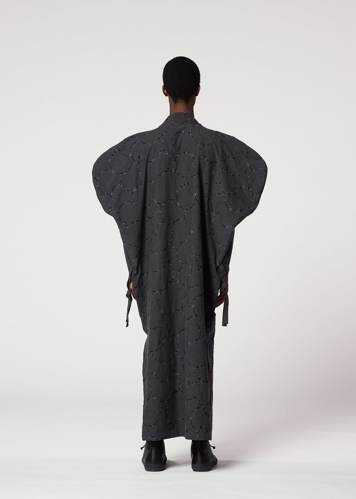 A person wearing the IM MEN VASE JACQUARD SHIRT, a long dark patterned piece with wide sleeves, stands facing away against a plain white background. Their arms are relaxed and they wear black shoes. view 6 of 7