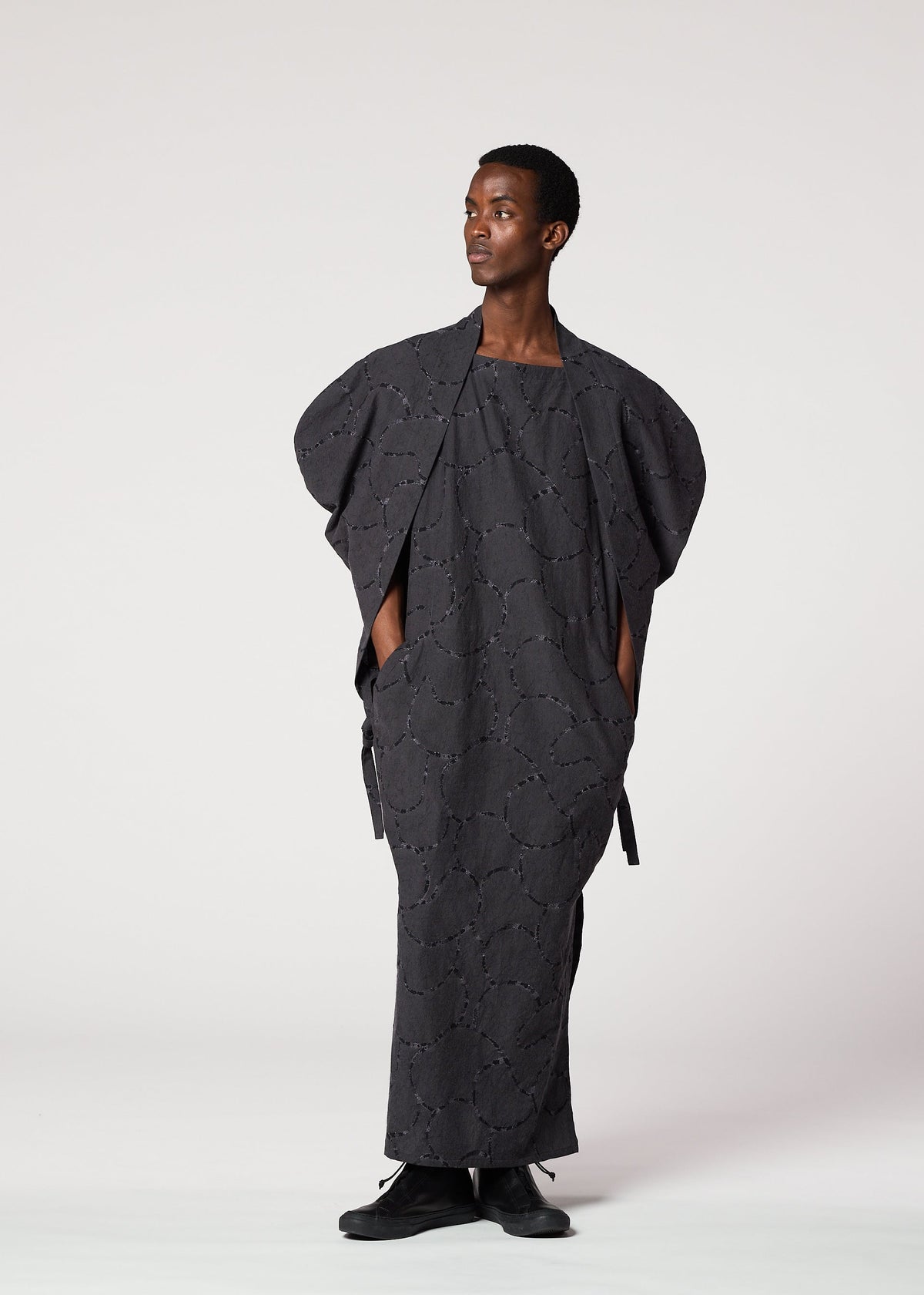 A person stands against a light background, wearing the IM MEN VASE JACQUARD SHIRT—a loose, dark gray long shirt with jacquard-inspired patterns, wide sleeves, and pockets—paired with black shoes and looking calmly to the side. view 7 of 7