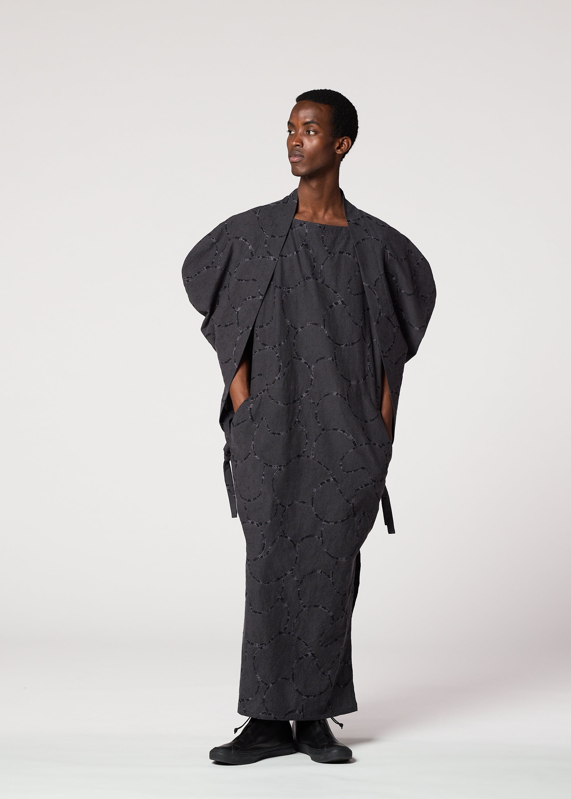 Zoomed view A person stands against a light background, wearing the IM MEN VASE JACQUARD SHIRT—a loose, dark gray long shirt with jacquard-inspired patterns, wide sleeves, and pockets—paired with black shoes and looking calmly to the side.