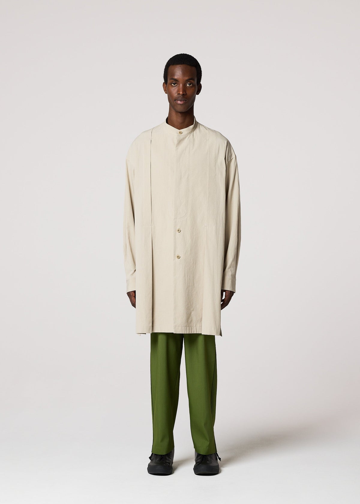 A person stands facing forward in the IM MEN WHITE SHIRT, a long button-up with asymmetrical details, paired with olive green pants and black shoes—an elegant outfit for formal events, set against a plain light background. view 5 of 8