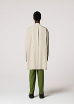 A person faces away, wearing the WHITE SHIRT by IM MEN—an elegant cream-colored cotton linen long shirt with a center pleat—paired with green pants and black shoes, ideal for formal occasions against a plain light background.