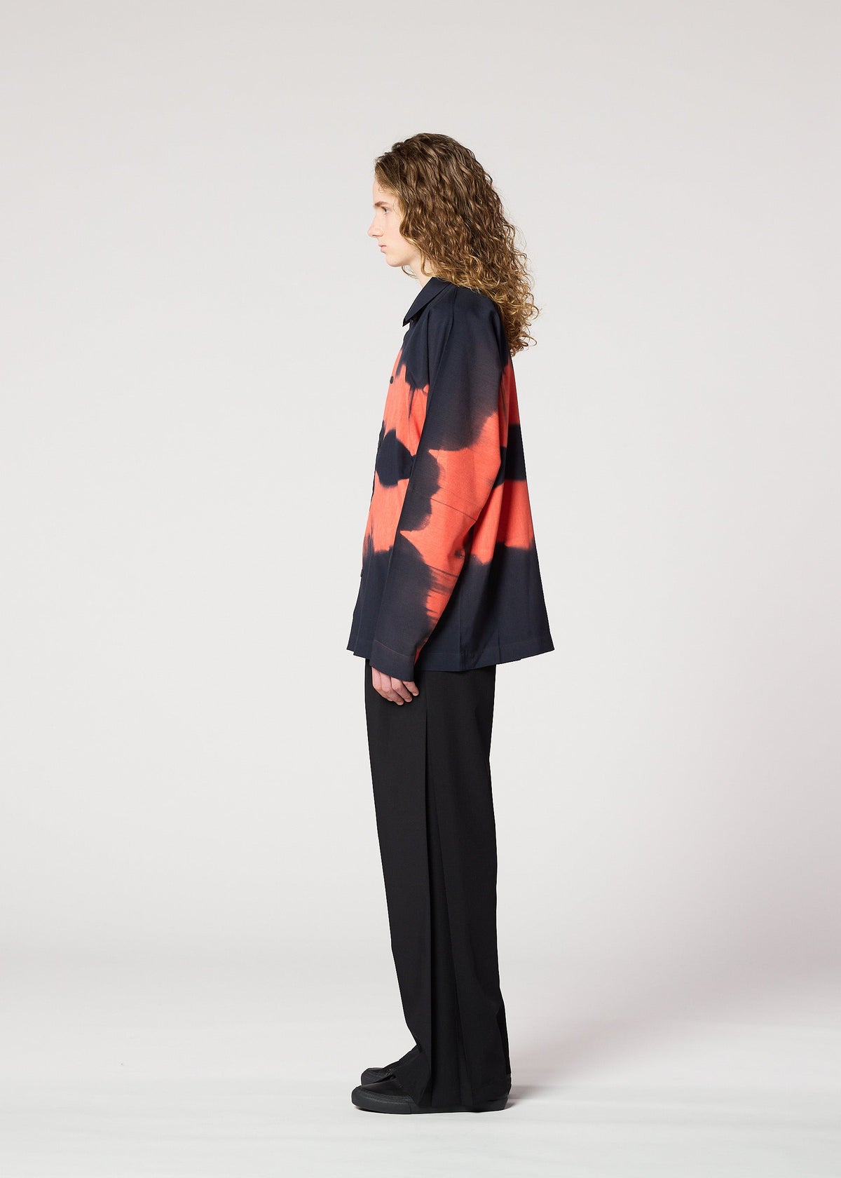 A person with curly hair stands in profile, wearing the IM MEN ITAJIME SHIRT with bold red and black geometric patterns, paired with black wide-leg pants and shoes, against a plain white background. view 7 of 9