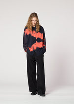 A person with long curly hair stands looking down, hands in pockets, wearing the loose-fitting black and red ITAJIME SHIRT by IM MEN with a subtle geometric pattern and wide-leg black pants against a plain light background.