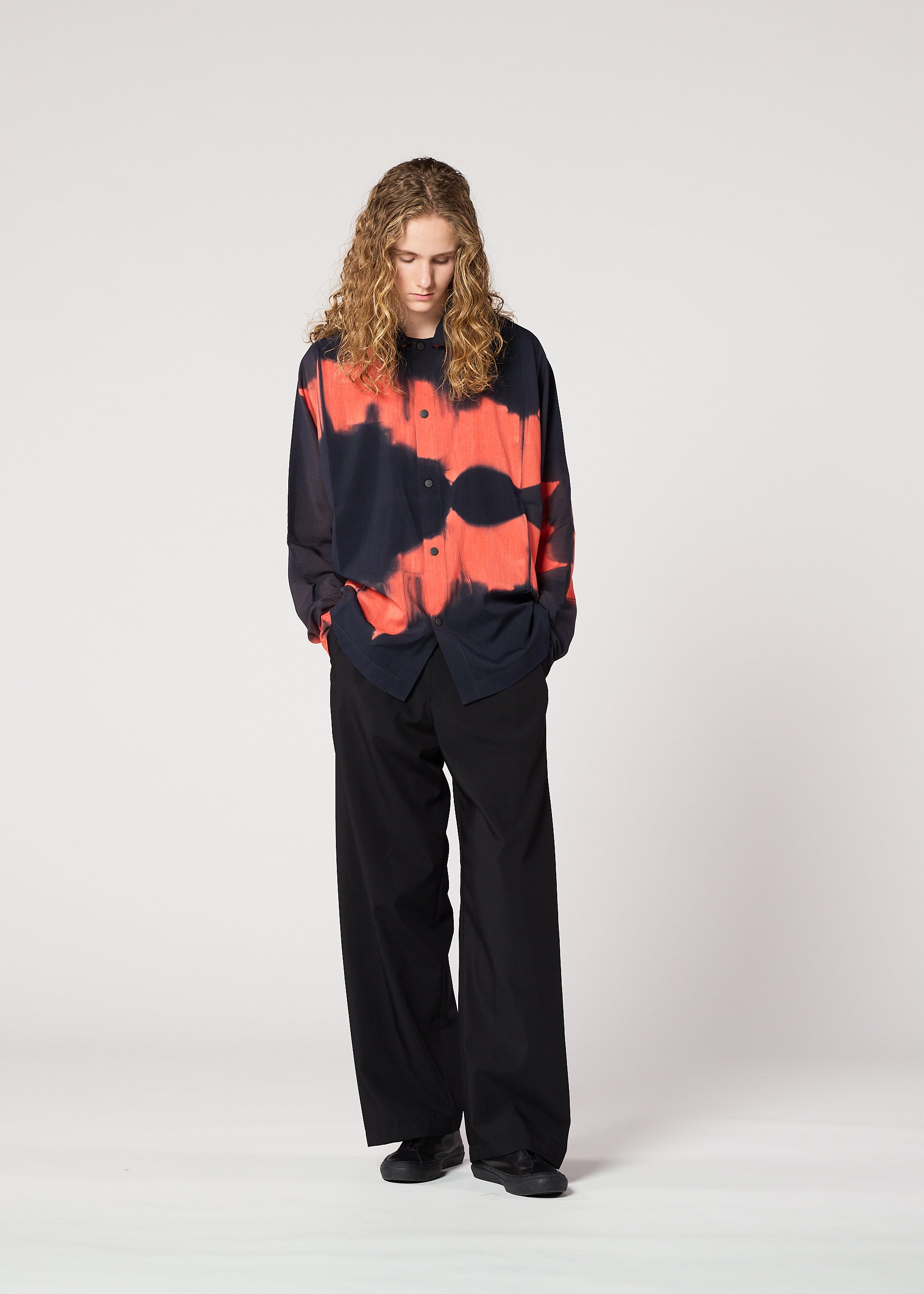 Zoomed view A person with long curly hair stands looking down, hands in pockets, wearing the loose-fitting black and red ITAJIME SHIRT by IM MEN with a subtle geometric pattern and wide-leg black pants against a plain light background.