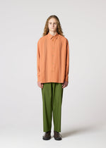 A person with long curly hair faces forward, wearing the IM MEN PLEATS SHIRT—an oversized peach pleated shirt made from recycled polyester—and loose green pants with dark shoes, against a plain light background.