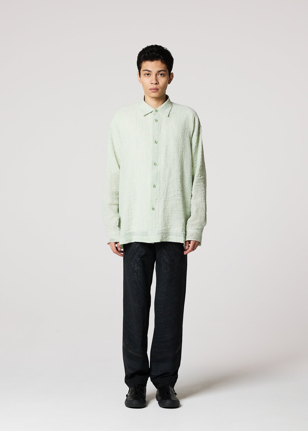 A person stands facing forward against a white background, wearing IM MEN’s STRIPE LINEN SHRINK light green shirt with loose black pants and black shoes. They have short dark hair and a neutral expression. view 6 of 9
