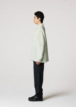 A young man stands in profile against a plain white background, wearing the IM MEN STRIPE LINEN SHRINK shirt in light green, paired with dark pants and black shoes.